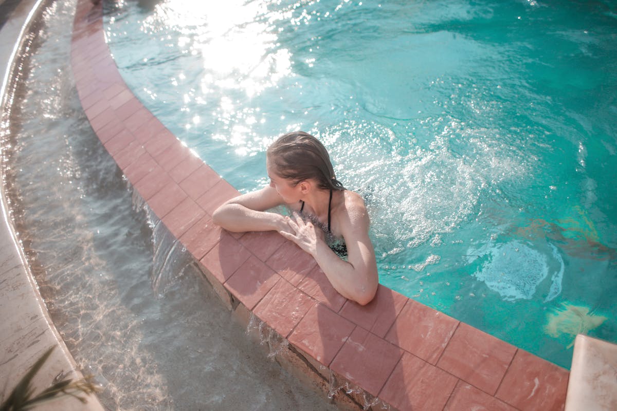 A woman enjoys a sunny day at the pool, leaning on the edge with tranquil waters.
