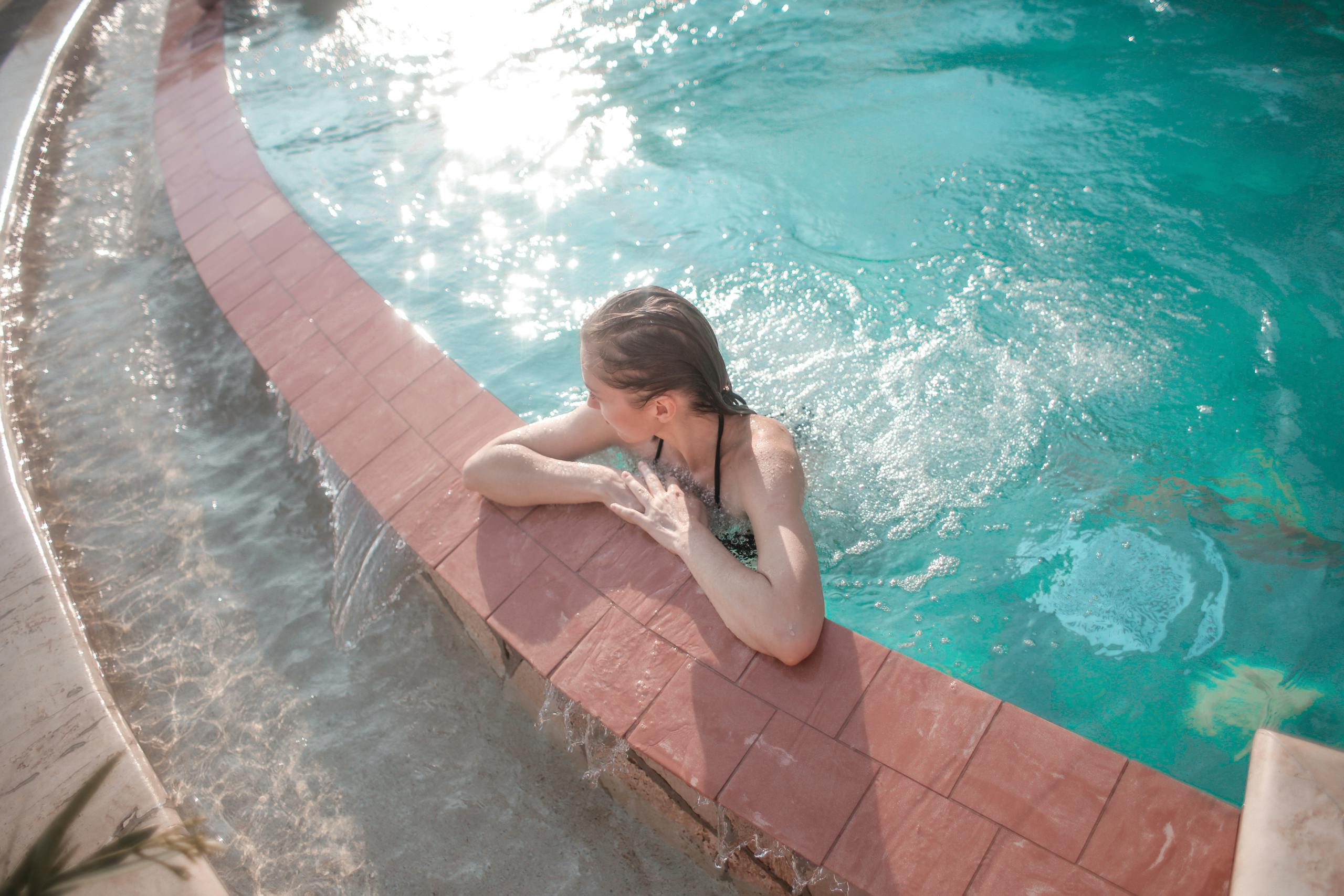 A woman enjoys a sunny day at the pool, leaning on the edge with tranquil waters.